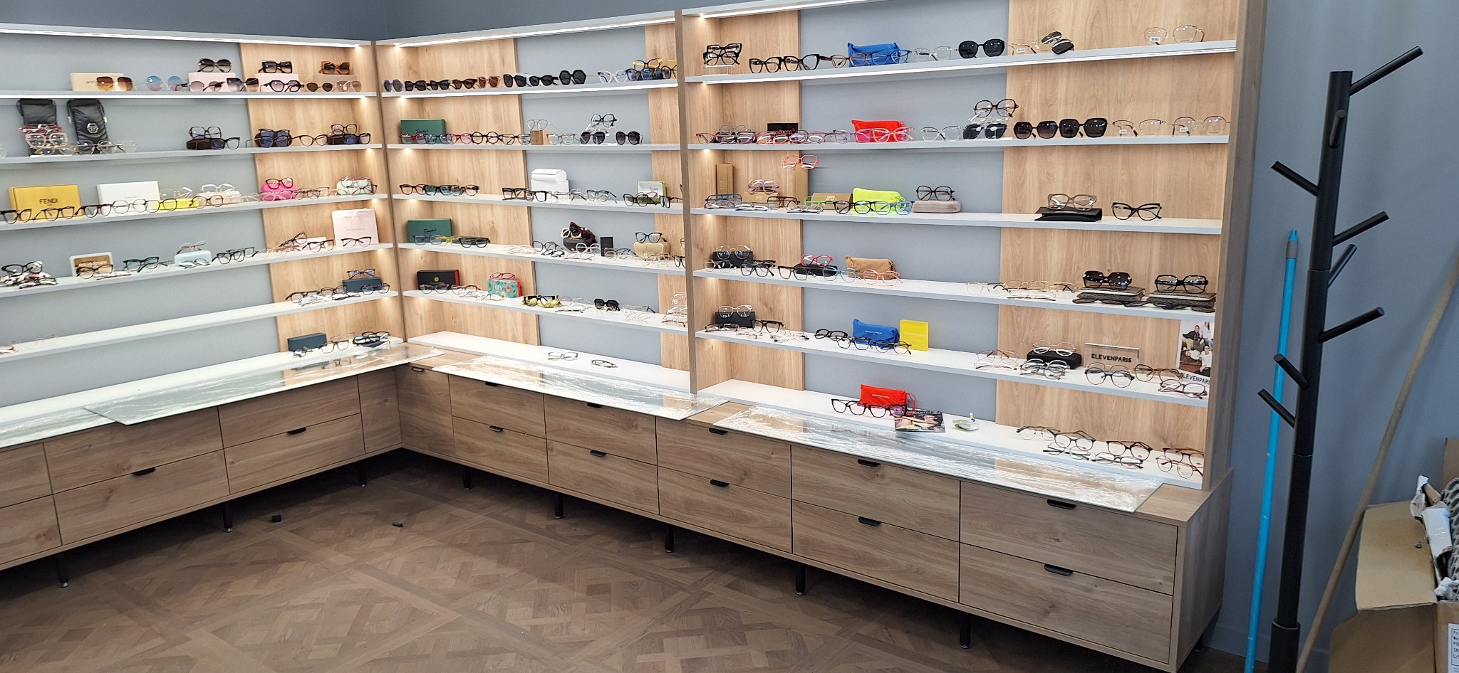 Wnętrze sklepu optycznego z drewnianymi szafkami i półkami, na których eksponowane są różnorodne okulary korekcyjne i przeciwsłoneczne.