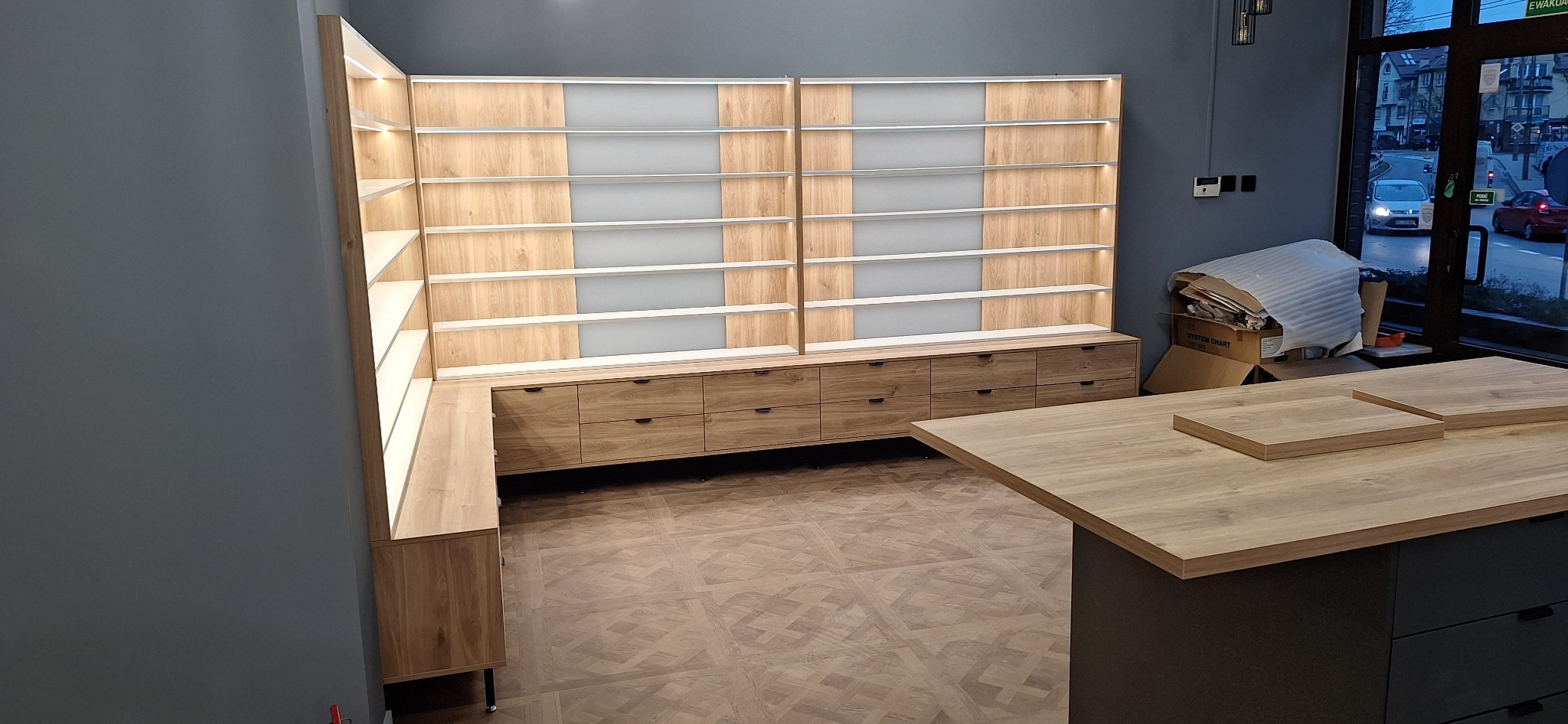 Duża, narożna szafa wnękowa z szufladami i podświetlanymi półkami, w jasnym odcieniu drewna, na podłodze wzór parkietu, widok na ulicę przez okno.