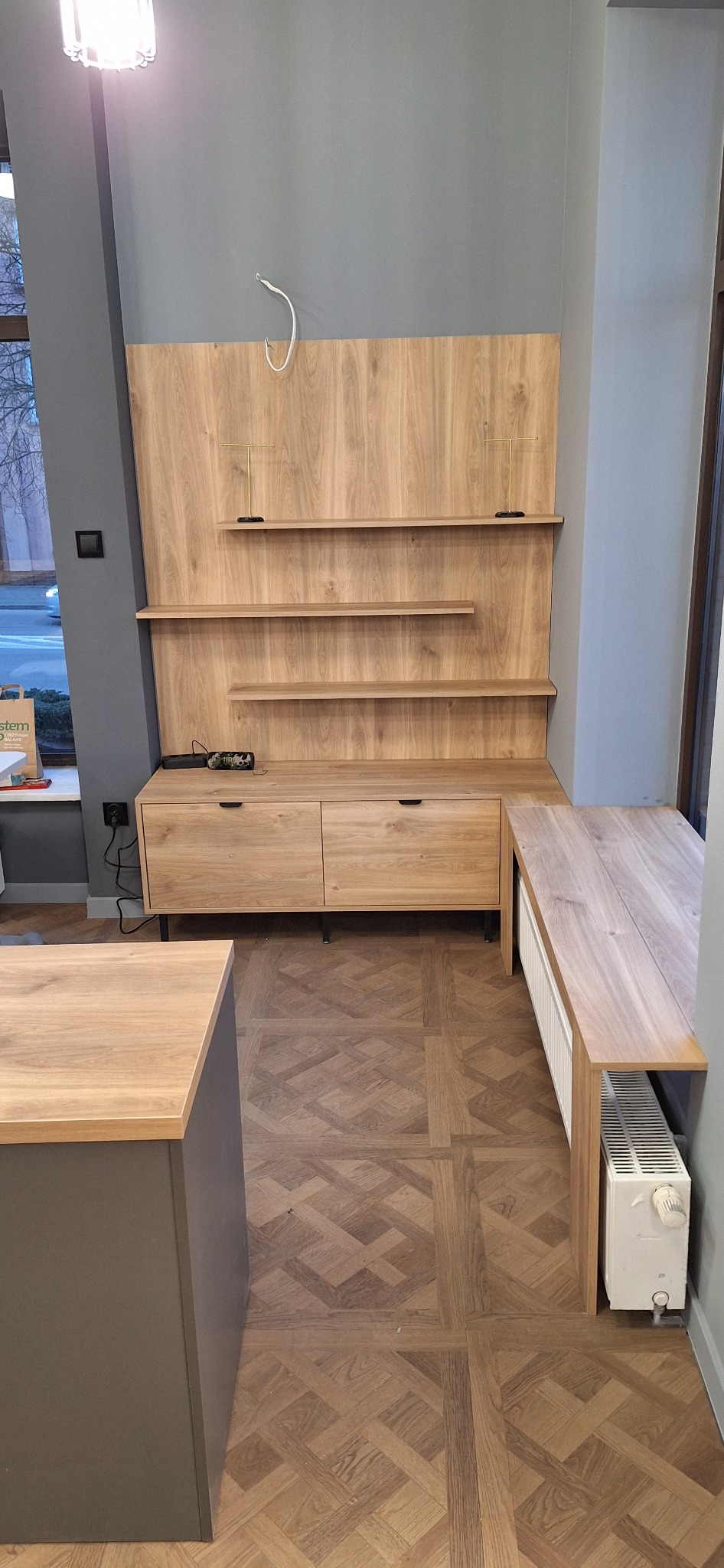 Drewniana zabudowa ściany z półkami i szafkami z szufladami, uzupełniona blatem roboczym przy oknie, na podłodze parkiet w jodełkę.