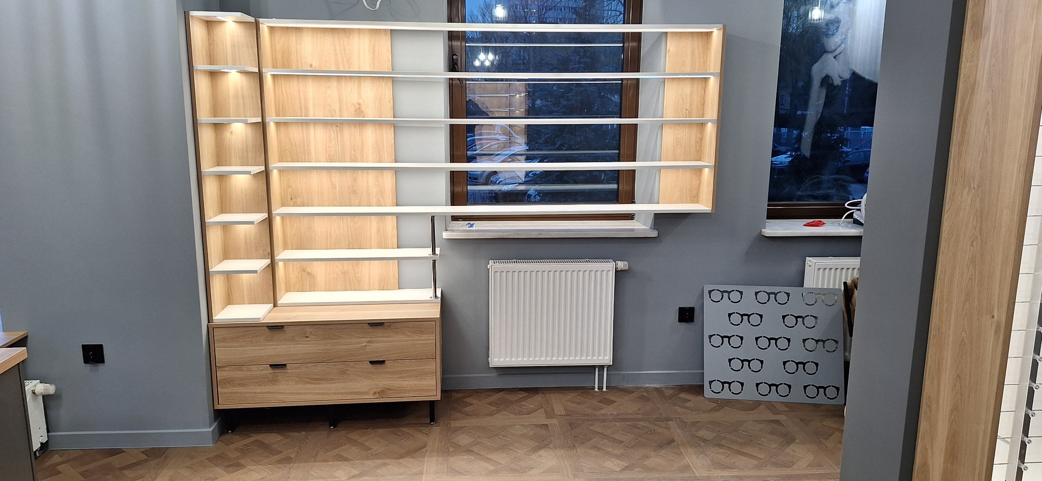 Drewniana szafa z półkami i szufladami, oświetlona od wewnątrz, ustawiona na tle okna i grzejnika, z dekoracyjną tablicą z motywem okularów obok.