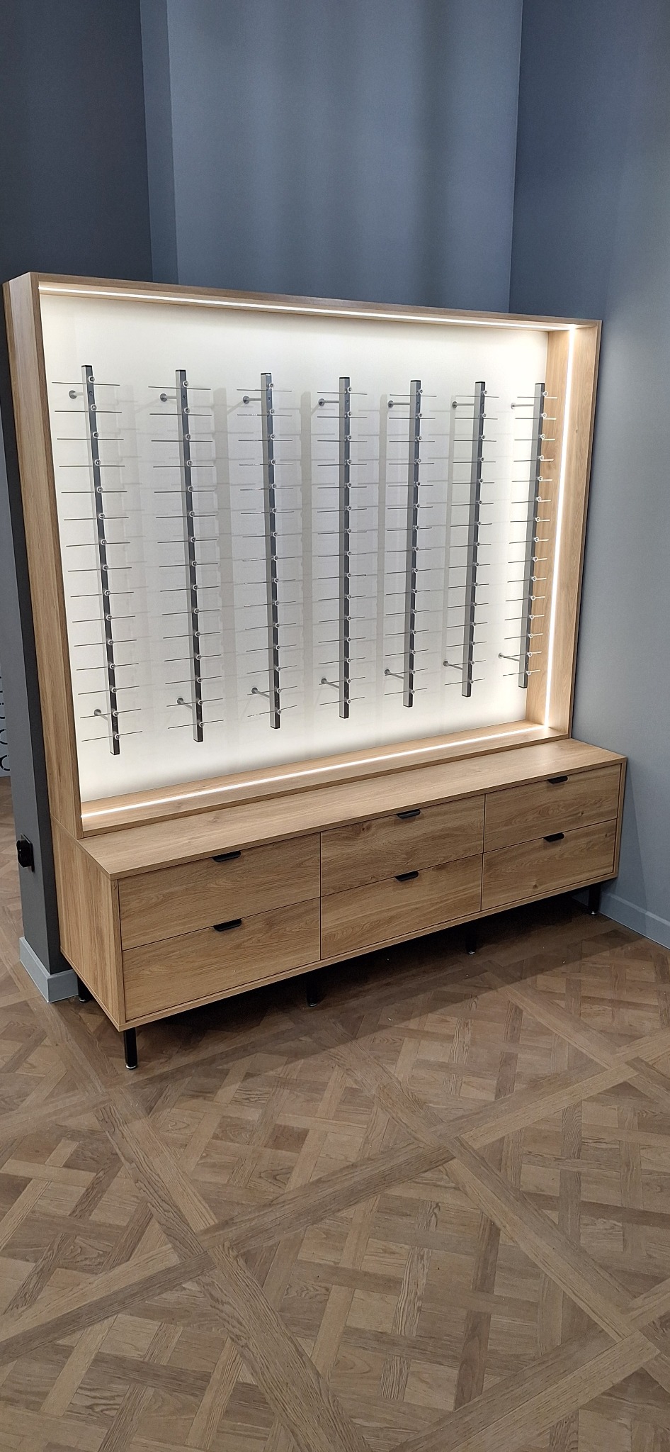 Ekspozytor okularów z podświetleniem LED, wykonany z jasnego drewna, zamontowany na komodzie z trzema szufladami, na tle szarej ściany i parkietu w geometryczny wzór.