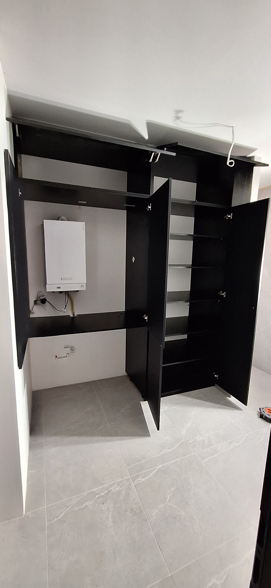 Czarna szafa na wymiar z otwartymi drzwiami, wnęka z piecem gazowym. Szare płytki na podłodze, białe ściany. Widoczne zawiasy i półki wewnątrz szafy.