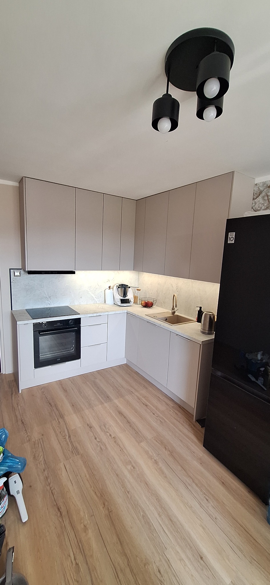 Jasna, narożna kuchnia na wymiar z beżowymi frontami, marmurowym blatem i czarnymi elementami AGD. Widoczna płyta indukcyjna, piekarnik i czarna lampa sufitowa.