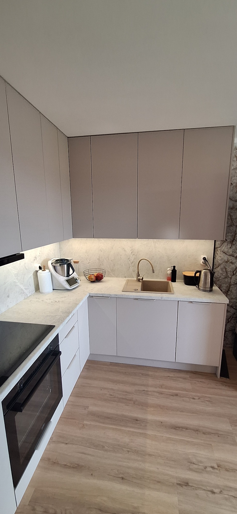 Jasna kuchnia z szarymi szafkami, marmurowym blatem i drewnianą podłogą. Na blacie widoczny robot kuchenny i owoce. Ciekawy wzór tapety na ścianie.