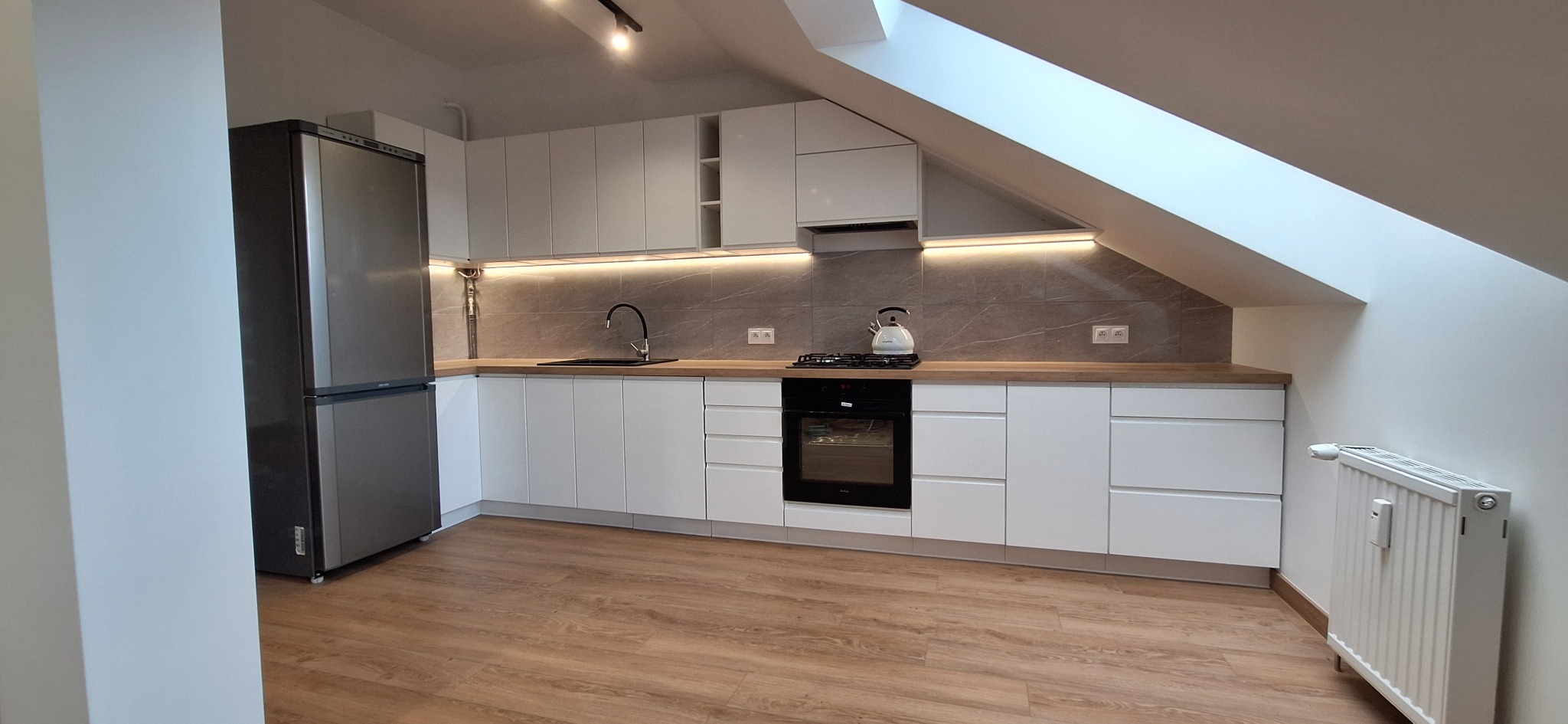 Biała kuchnia na poddaszu ze skośnym oknem dachowym i szarą lodówką. Minimalistyczny styl, oświetlenie LED pod szafkami. Płyta grzewcza z czajnikiem.