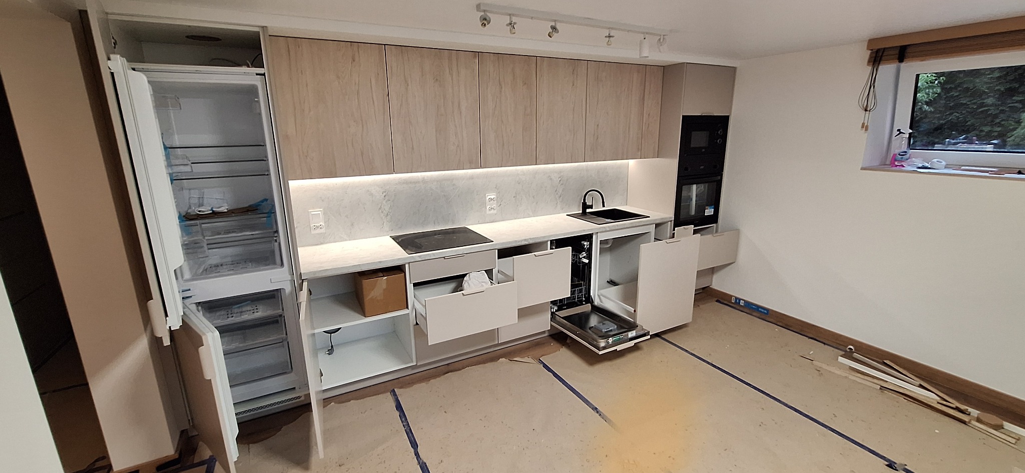 Jasna kuchnia z drewnopodobnymi szafkami i otwartymi szufladami, widoczne niedokończone wykończenie podłogi. Oświetlenie podszafkowe podkreśla blat.