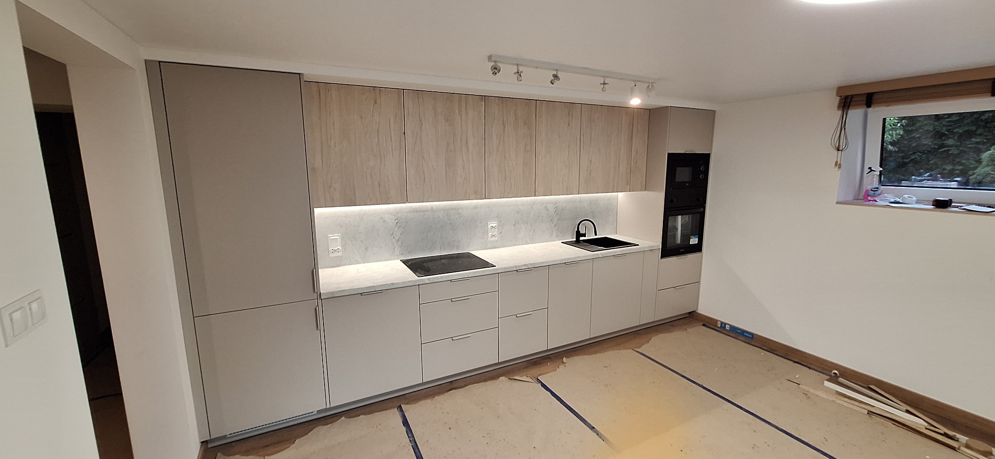 Jasna kuchnia z drewnianymi szafkami górnymi i szarymi dolnymi, podświetlony blat, czarny zlew i płyta grzewcza, okno z roletą.