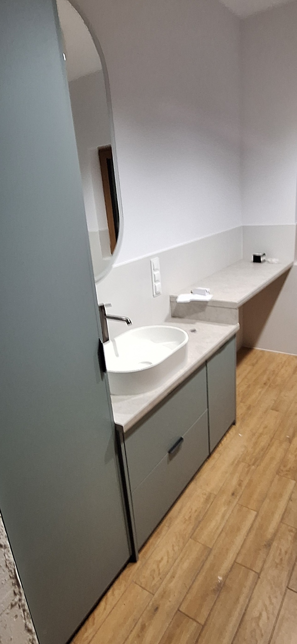 Nowoczesna łazienka z owalną umywalką nablatową i szarą szafką z szufladami. Uwagę zwraca duże, owalne lustro i drewnopodobna podłoga. Minimalistyczny design.