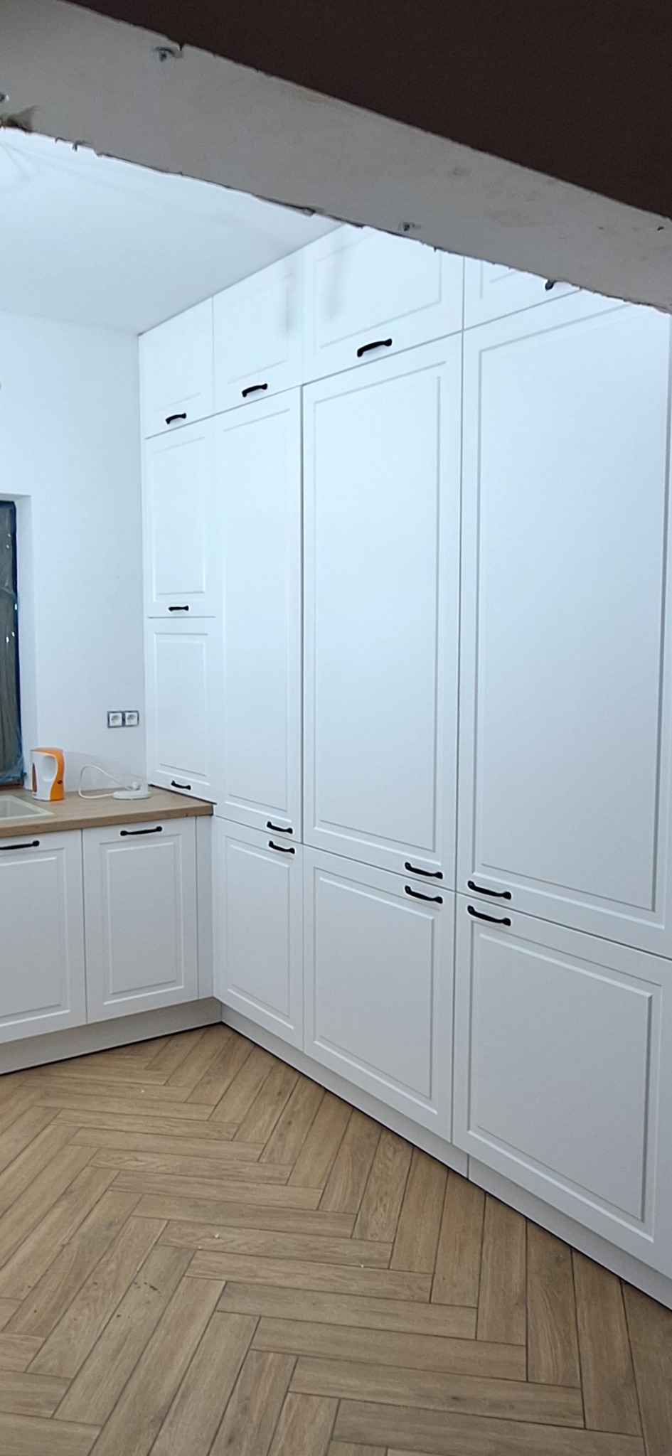 Biała zabudowa kuchenna z czarnymi uchwytami, parkiet w jodełkę. Widoczne szafki dolne i górne, blat drewniany. Minimalistyczny design, nowoczesne wykończenie.