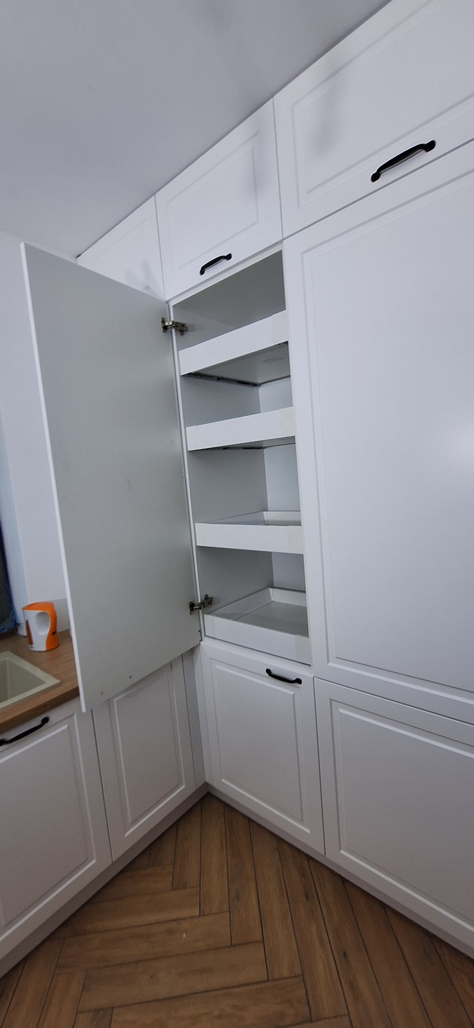 Biała zabudowa kuchenna z otwartą szafką cargo, widoczne półki. Ujęcie z perspektywy podłogi, na której ułożono parkiet w jodełkę. Minimalistyczny design.