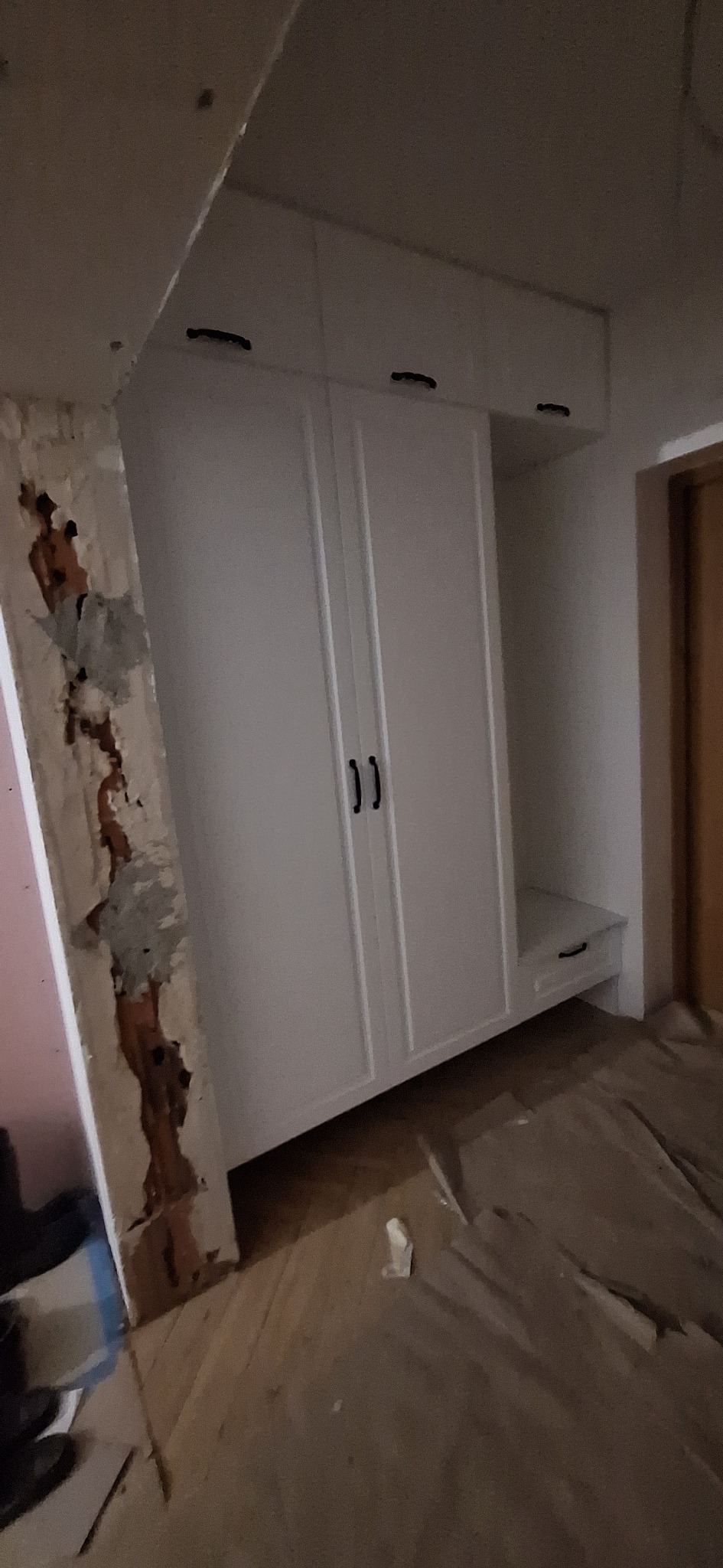 Biała szafa wnękowa z czarnymi uchwytami, widoczna na tle remontowanego pomieszczenia z odsłoniętą ścianą i drewnianą podłogą. Ujęcie z dołu.