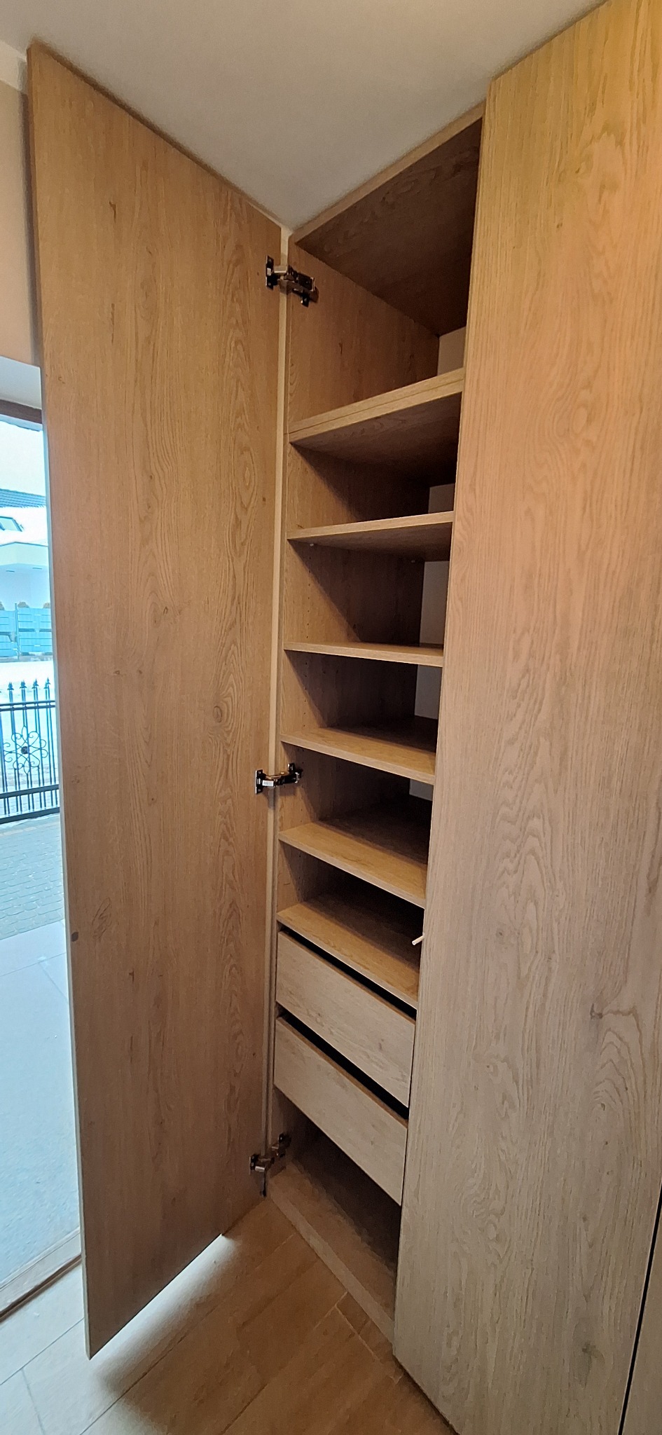 Otwarta, jasna szafa z drewnianymi półkami i szufladami, widoczne zawiasy, na tle okno z widokiem na zewnątrz. Naturalne wykończenie drewna.