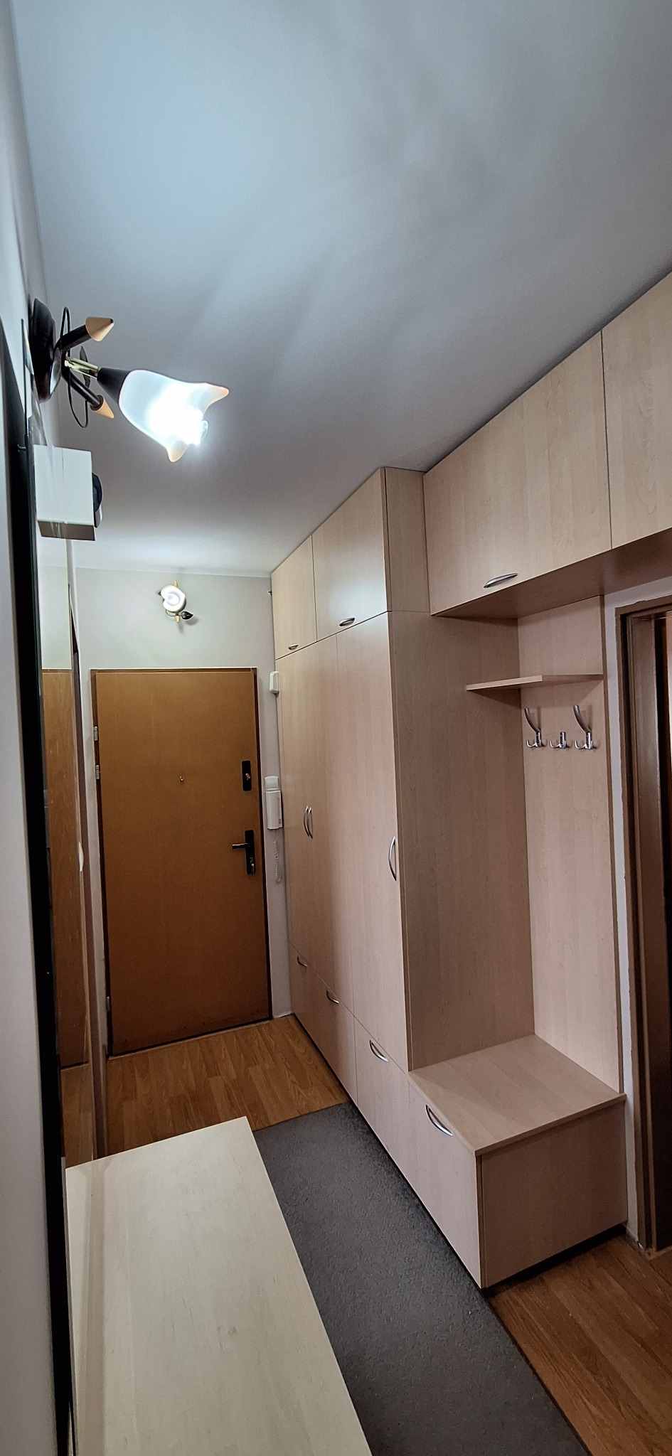 Jasna zabudowa przedpokoju z szafami, siedziskiem i wieszakami, oświetlona lampą w stylu retro. Funkcjonalne rozwiązanie do przechowywania w wąskim wnętrzu.