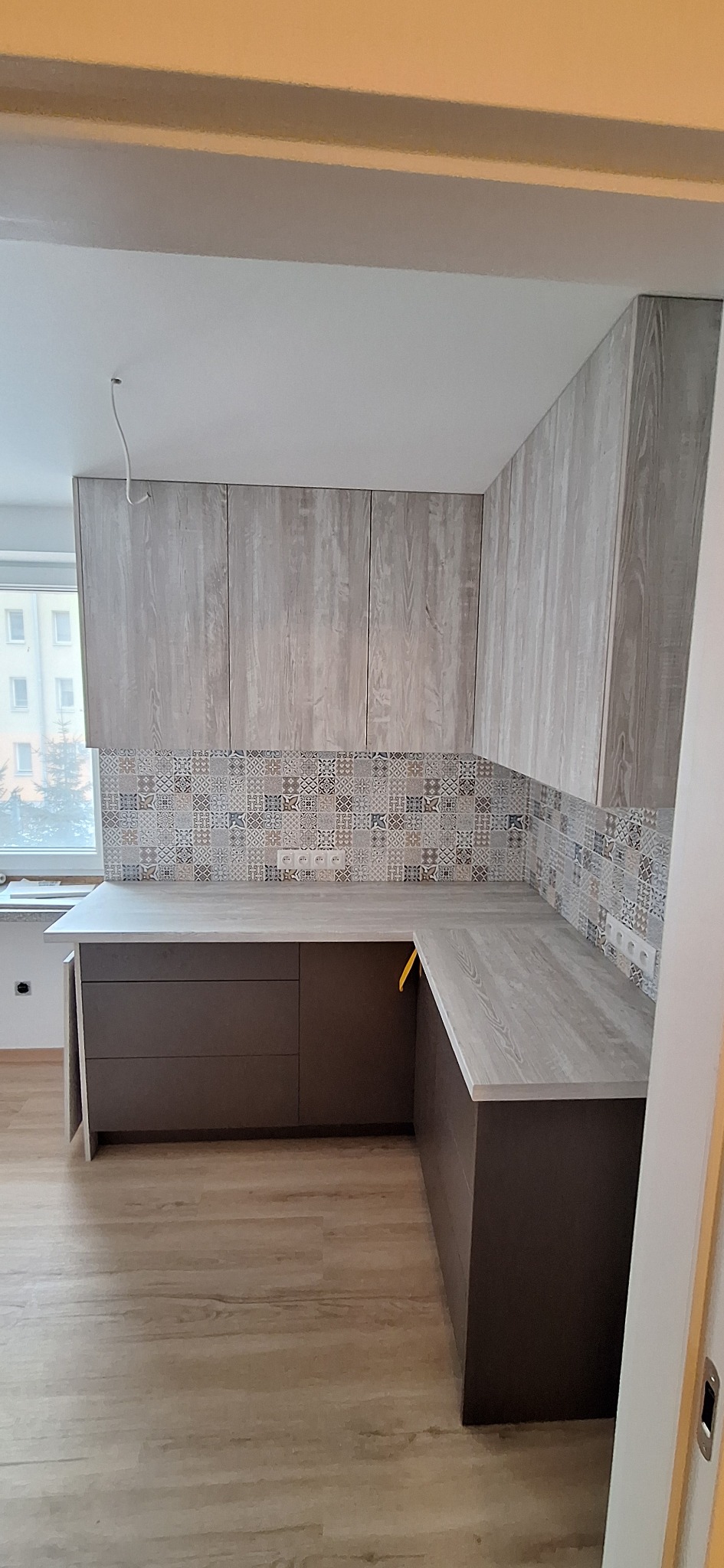 Jasna kuchnia z drewnianymi szafkami i wzorzystymi płytkami nad blatem. Ujęcie z perspektywy wejścia, widoczna podłoga i fragment okna z widokiem na budynek.