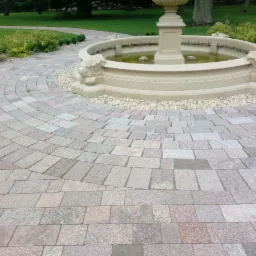 Okrągła ścieżka z szarej kostki prowadząca do fontanny w ogrodzie, widoczne nieregularne ułożenie kostki i małe kamyczki wokół fontanny.
