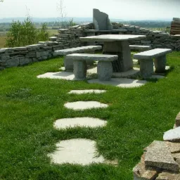 Ogród z kamiennym stołem i ławkami, ścieżka z jasnych płyt prowadzi przez trawnik, otoczony niskim murem z kamienia łupanego, w tle zielone pola i lasy.