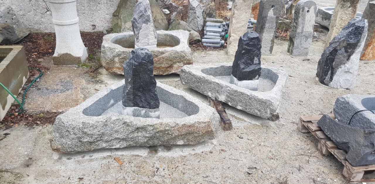 Granitowe poidełka dla ptaków z ciemnymi obeliskami wewnątrz, ustawione na żwirowym podłożu.