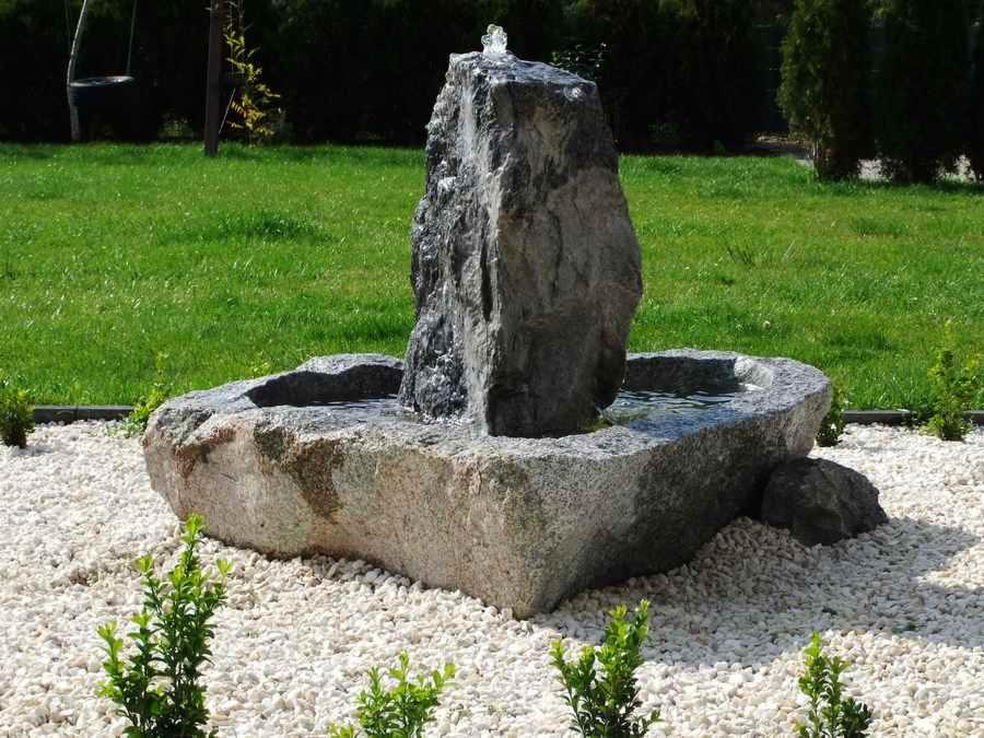 Ozdobna fontanna ogrodowa z szarego kamienia, otoczona białym żwirem i niską zielenią, na tle trawnika.