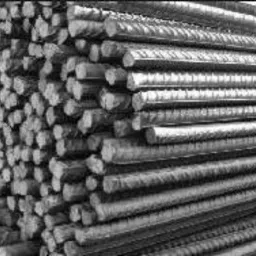 Stos żebrowanych prętów z metalu, ułożonych równolegle i ciasno obok siebie. Widoczne przekroje poprzeczne prętów. Zdjęcie czarno-białe.