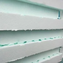 Bliski kadr na stos płyt izolacyjnych z białego styropianu z widocznymi zielonymi krawędziami i szczelinami między nimi, ukazującymi strukturę materiału.