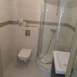 Jasna łazienka po remoncie z kabiną prysznicową narożną, wiszącą toaletą i umywalką z szafką w odcieniach brązu, wykończona białymi płytkami z dekoracyjnym pasem mozaiki.