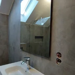 Nowoczesna łazienka z szarymi płytkami, białą umywalką z szafką, lustrem odbijającym okno dachowe i widocznymi otworami na baterię.