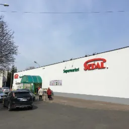 Biała elewacja budynku supermarketu z czerwonym logo Sedal i zielonym napisem Supermarket, widoczne osoby wchodzące do sklepu, zaparkowane samochody, drzewa bez liści i błękitne niebo.
