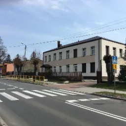 Dwukondygnacyjny budynek z jasną elewacją, częściowo ocieplony, widoczny z perspektywy ulicy z przejściem dla pieszych i żółtymi barierkami.