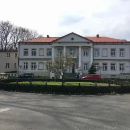 Dwupiętrowy budynek z kolumnami i czerwonym dachem, widoczny z ulicy z okrągłym klombem, kilka zaparkowanych samochodów, drzewa bez liści.