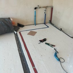 HYDRO-THERM SPÓŁKA Z OGRANICZONĄ ODPOWIEDZIALNOŚCIĄ - Rozłożona izolacja pod ogrzewanie podłogowe z widocznymi rurami, narzędziami i elementami instalacji. W tle surowe ściany w trakcie prac wykończeniowych.