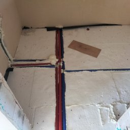HYDRO-THERM SPÓŁKA Z OGRANICZONĄ ODPOWIEDZIALNOŚCIĄ - Izolacja podłogowa z rurami grzewczymi w kolorze czerwonym i niebieskim, zabezpieczonymi pianką montażową. Widoczna płyta drewniana na izolacji.