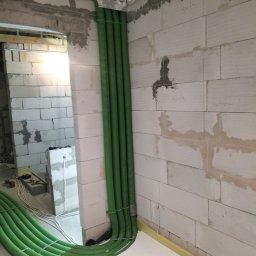 HYDRO-THERM SPÓŁKA Z OGRANICZONĄ ODPOWIEDZIALNOŚCIĄ - Zielone rury instalacyjne, prowadzone wzdłuż surowej ściany z betonu komórkowego, zabezpieczone srebrną folią. Widoczne mocowania i fragment podłogi z izolacją.