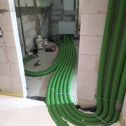 HYDRO-THERM SPÓŁKA Z OGRANICZONĄ ODPOWIEDZIALNOŚCIĄ - Zielone, karbowane rury instalacyjne ułożone wzdłuż ściany z białych bloczków, prowadzące do wnętrza budynku w trakcie budowy. Widoczne narzędzia i materiały budowlane.