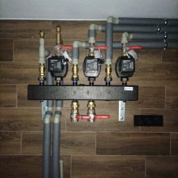 HYDRO-THERM SPÓŁKA Z OGRANICZONĄ ODPOWIEDZIALNOŚCIĄ - Rozdzielacz ogrzewania podłogowego z trzema pompami obiegowymi, zamontowany na ścianie z brązowych płytek imitujących drewno. Rury izolowane szarą pianką.