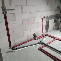 HYDRO-THERM SPÓŁKA Z OGRANICZONĄ ODPOWIEDZIALNOŚCIĄ - Czerwone i szare rury instalacji wodnej/kanalizacyjnej ułożone na surowym betonie w pomieszczeniu z białymi ścianami z betonu komórkowego. Widoczna drabina.