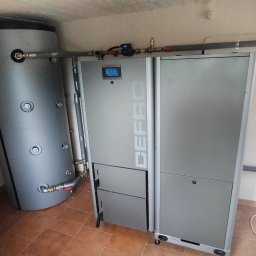 HYDRO-THERM SPÓŁKA Z OGRANICZONĄ ODPOWIEDZIALNOŚCIĄ - Szary piec CO marki DEFRO z podłączonym zasobnikiem wody, widoczne rury i armatura. Całość w pomieszczeniu z brązową terakotą.