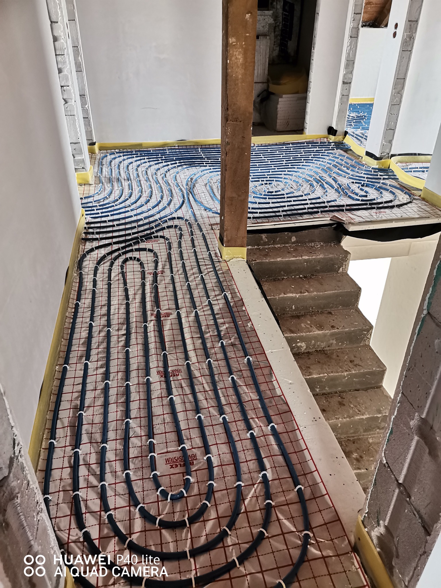 Niebieskie rurki ogrzewania podłogowego ułożone na folii z siatką, widoczne fragmenty surowych ścian i schodów w trakcie remontu.