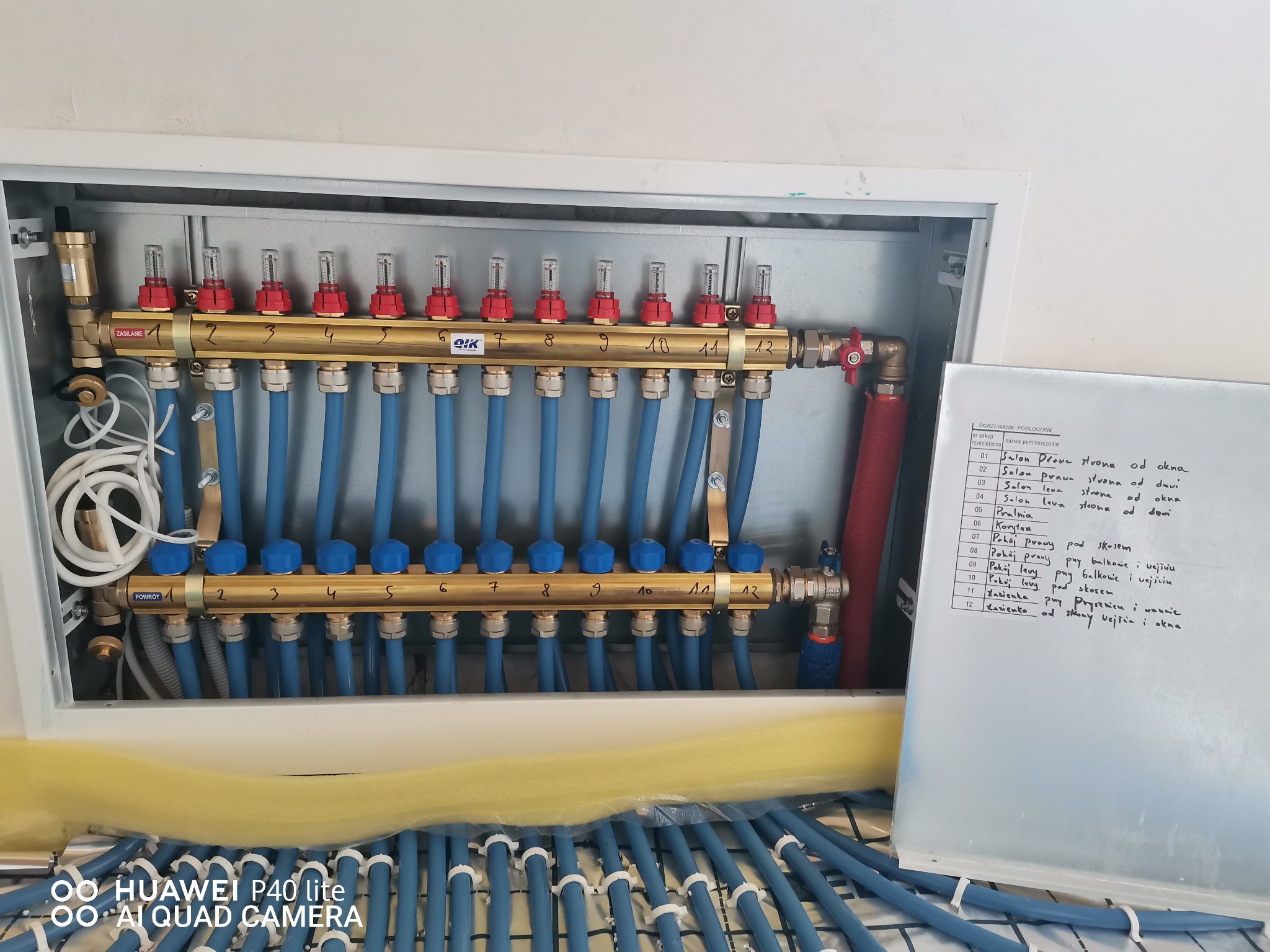 Rozdzielacz ogrzewania podłogowego z rurami w szafce, widoczna lista pomieszczeń z przypisanymi numerami sekcji, żółta pianka izolacyjna.