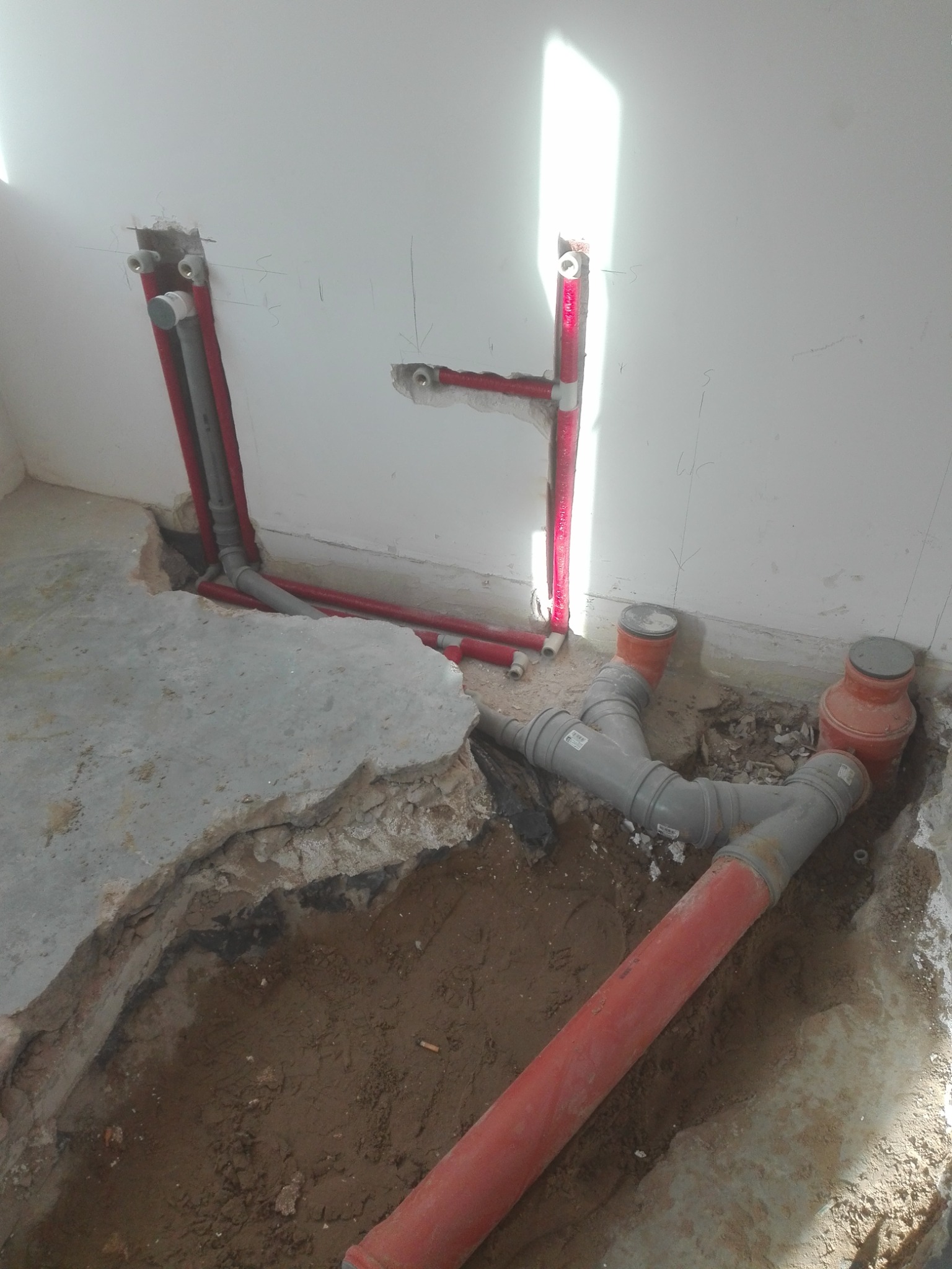 Wykopane rury kanalizacyjne i instalacyjne w trakcie montażu w pomieszczeniu z surowym betonem i ścianami. Widoczne połączenia trójnikowe i pionowe podejścia do ściany.