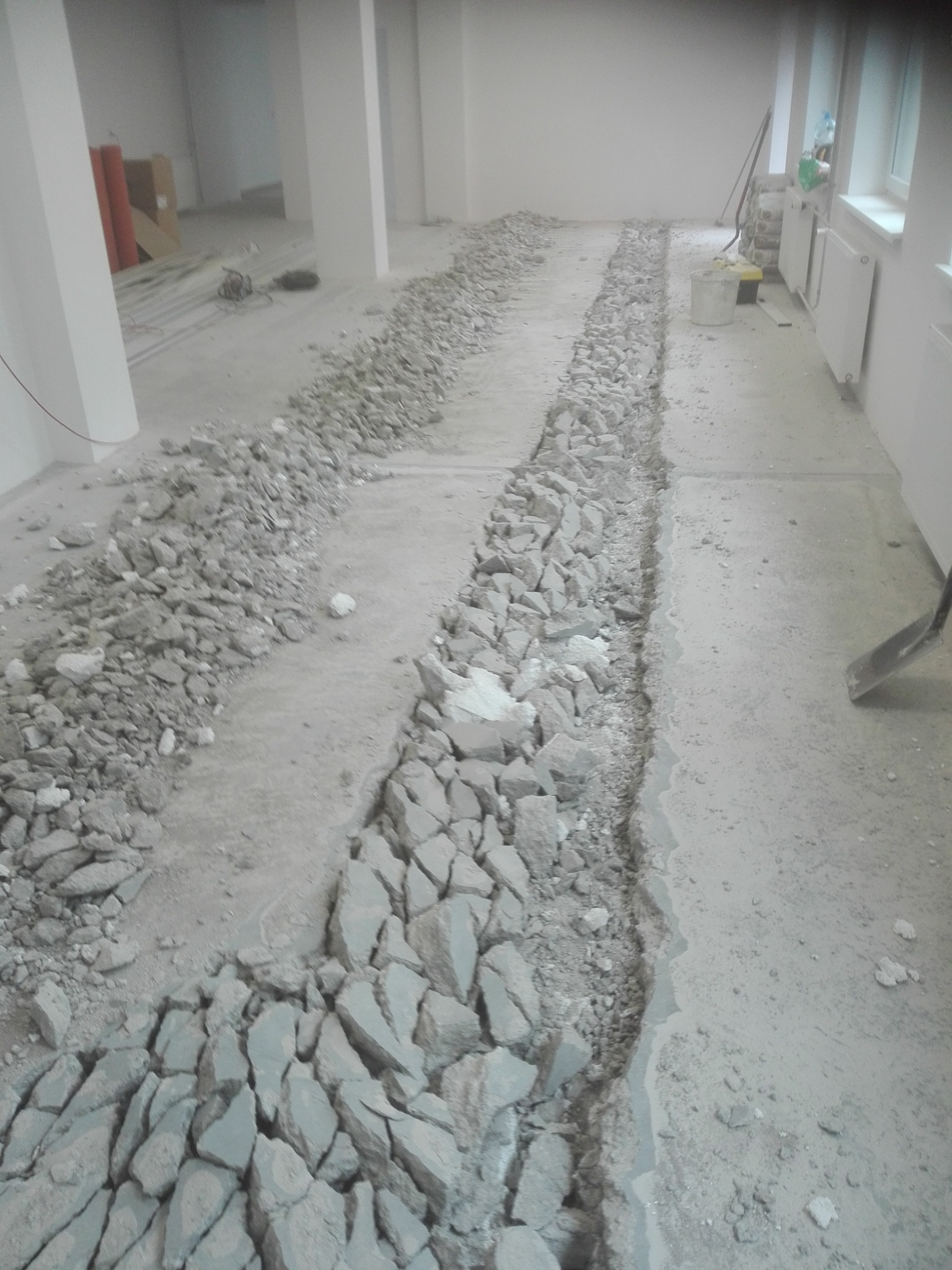 Wykuta bruzda w betonowej posadzce pod instalację, widoczne gruz i narzędzia w tle, wnętrze w trakcie remontu.