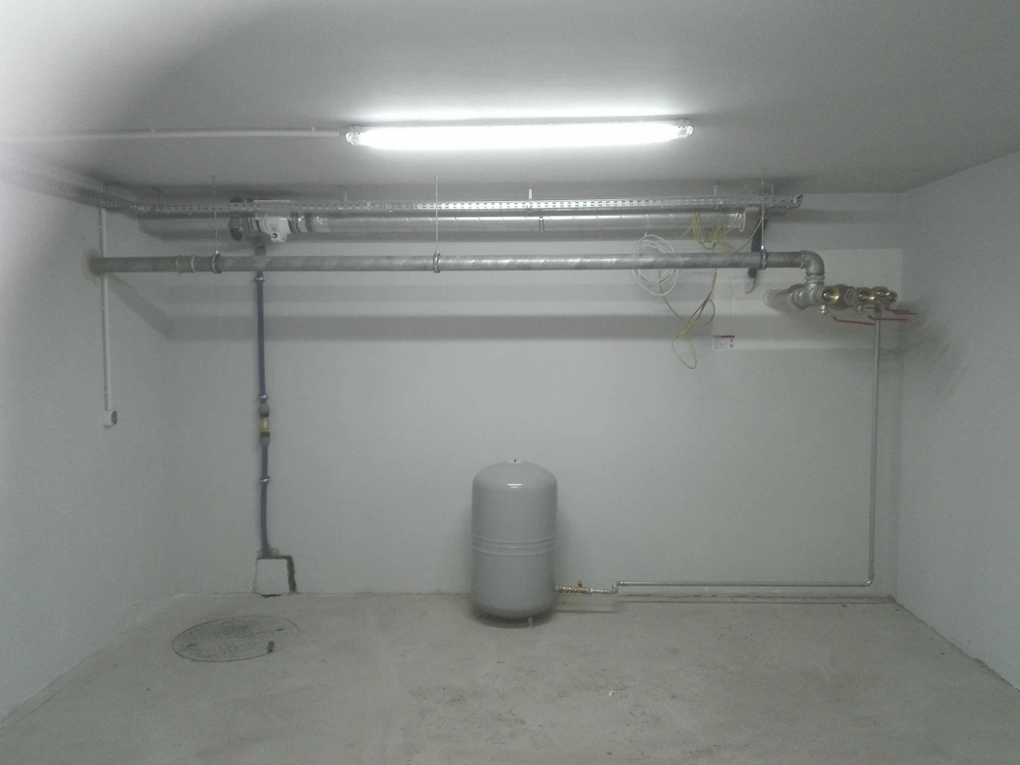 Szara instalacja wodna z zbiornikiem ciśnieniowym w piwnicy w Golub-Dobrzyń, widoczne rury na ścianach i suficie, oświetlenie jarzeniowe.