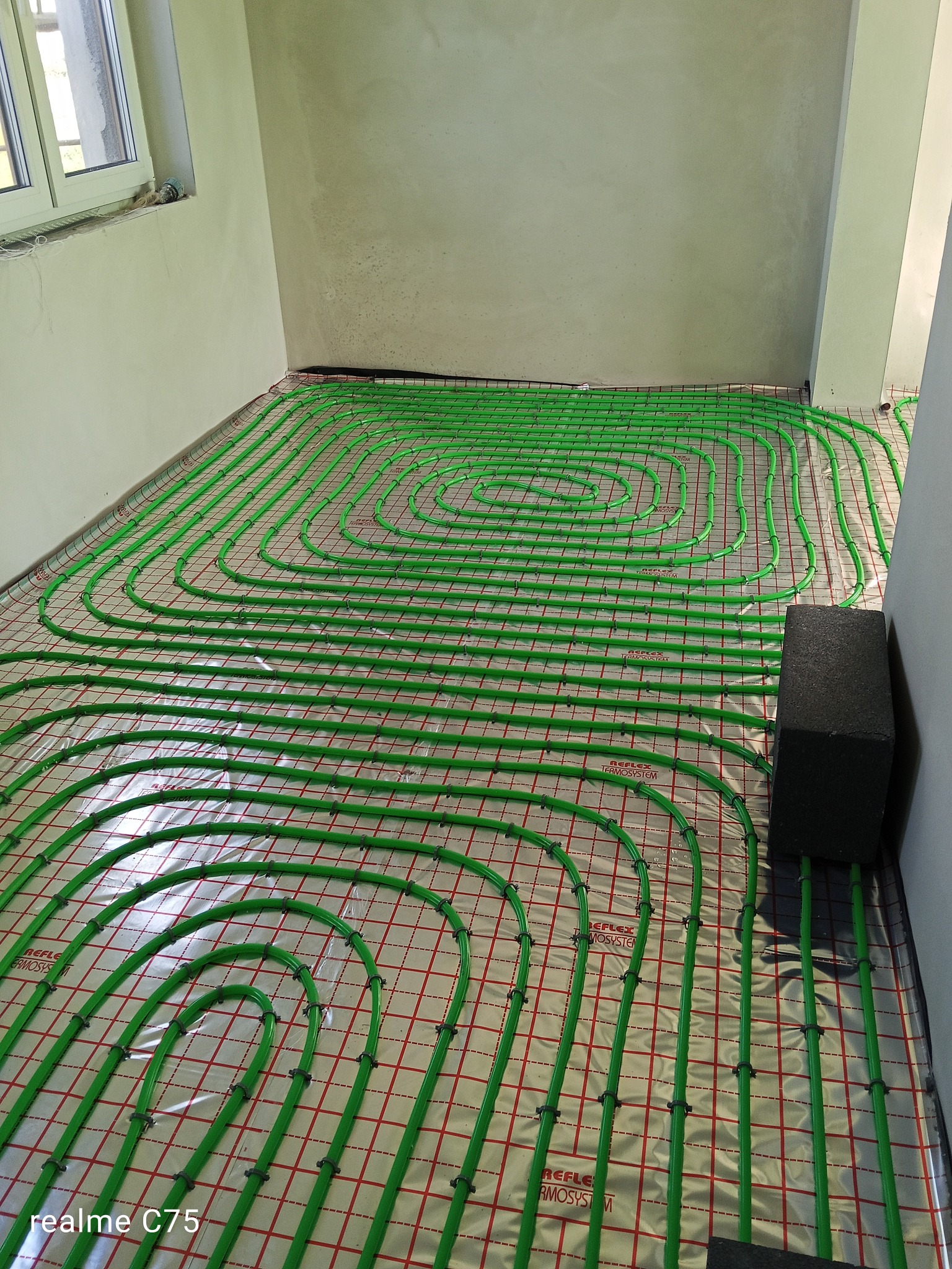 Zielone rury ogrzewania podłogowego ułożone spiralnie na folii z czerwoną siatką, widoczne czarne uchwyty mocujące i fragment ściany w surowym stanie.