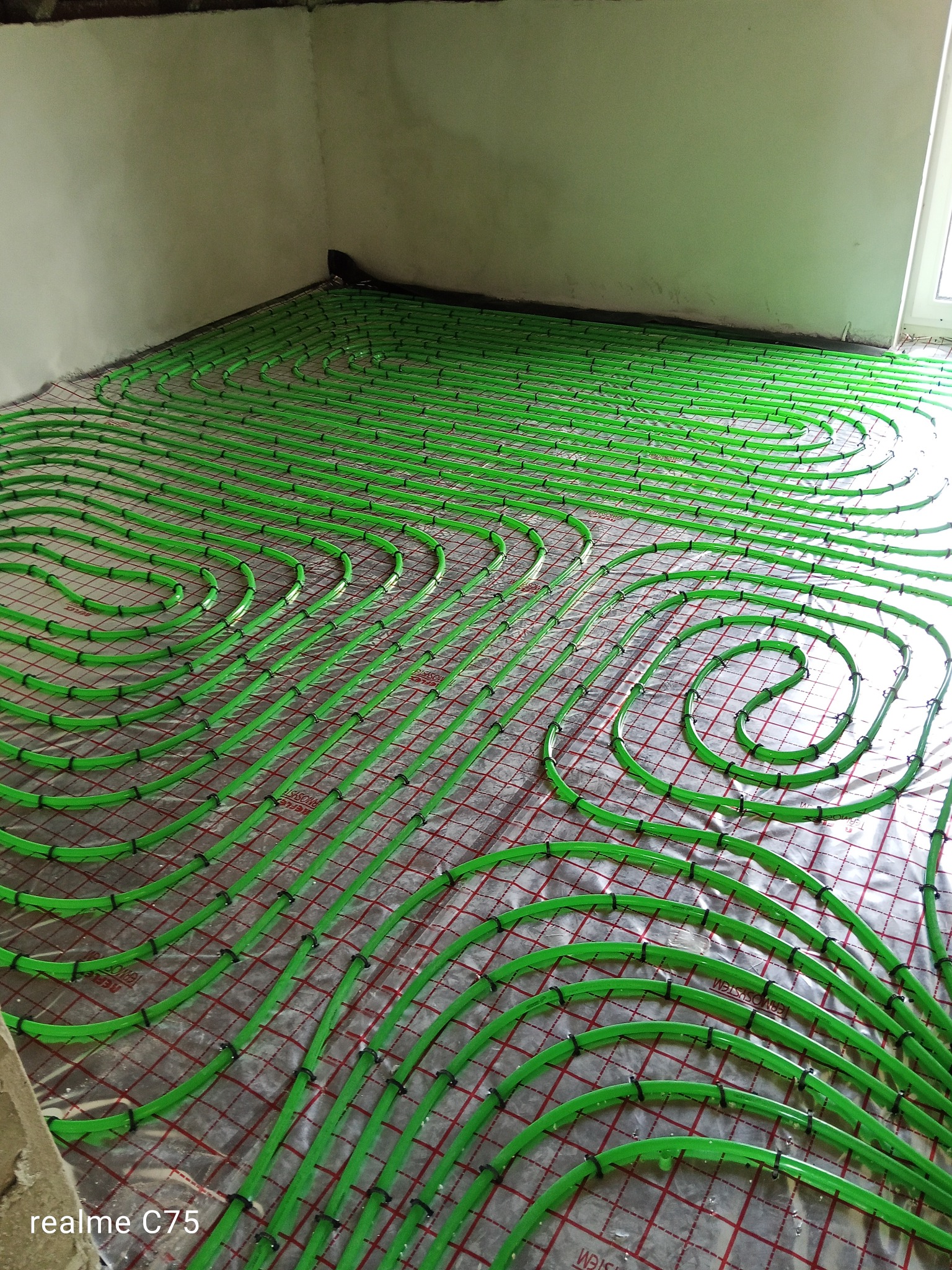 Zielone rury ogrzewania podłogowego ułożone na czerwonej siatce w jasnym pomieszczeniu. Widoczne czarne mocowania rur. Ujęcie z góry, ukośnie.