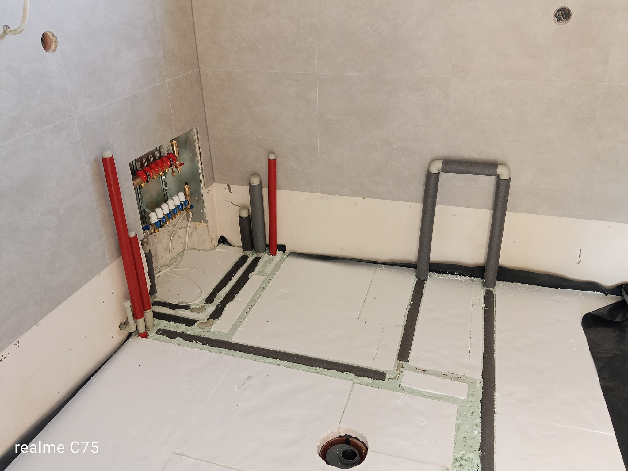 Biała izolacja podłogowa z czarnymi taśmami i rurami grzewczymi w łazience z szarą płytką. Widoczny rozdzielacz i odpływ podłogowy.