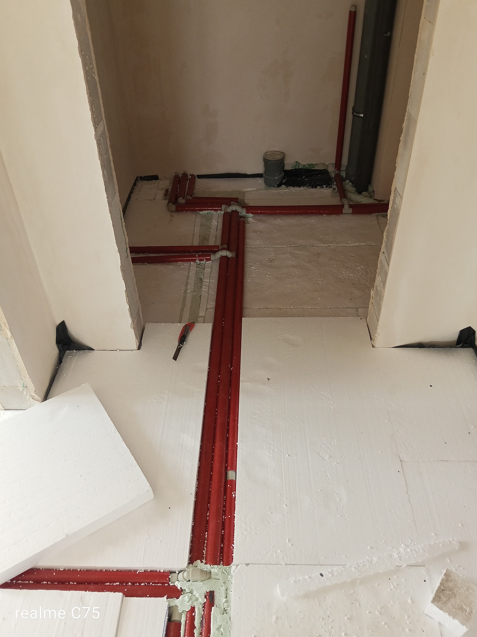 Czerwone rury instalacji wod-kan ułożone na białym styropianie w trakcie montażu. Widoczne narzędzie do cięcia styropianu i fragment ściany w surowym stanie.