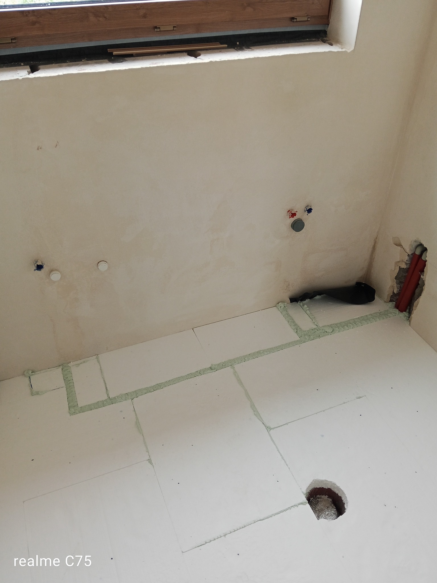 Przygotowanie do ogrzewania podłogowego: białe płyty izolacyjne z pianką montażową na podłodze, widoczne rury w ścianie i otwory na instalacje. Surowy stan, okno w tle.