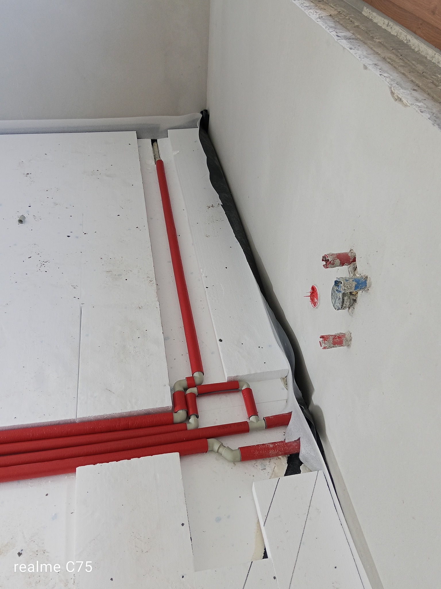 Czerwone rury ogrzewania podłogowego ułożone na białym styropianie, widoczne przyłącza do ściany. Etap prac przed zalaniem posadzki.
