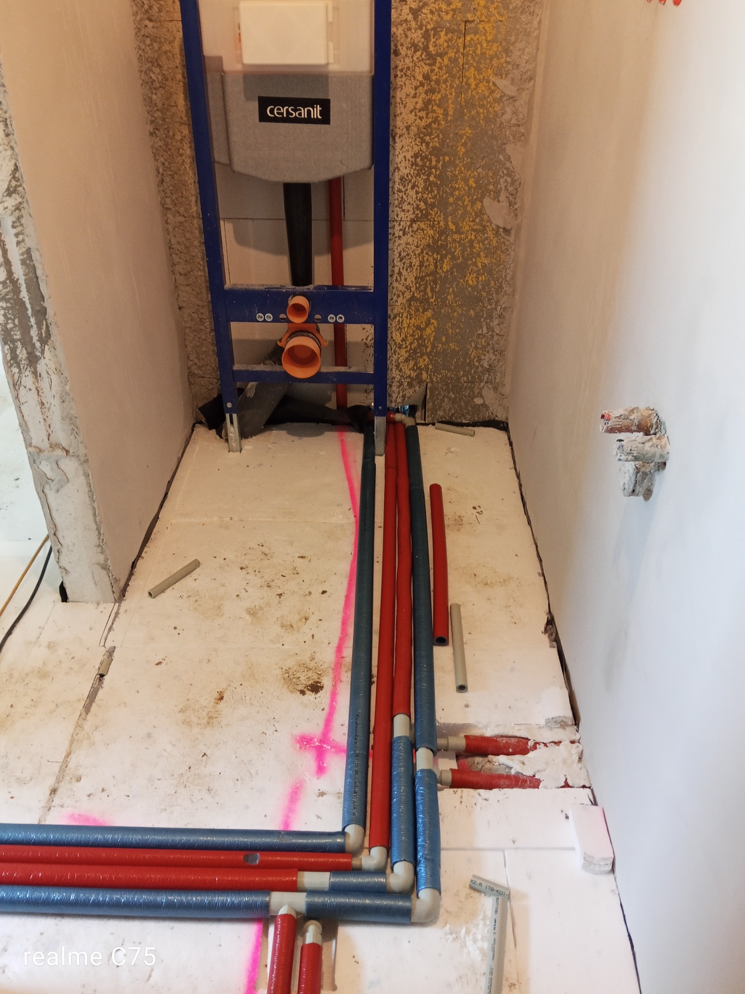 Instalacja ogrzewania podłogowego w łazience: rury grzewcze w izolacji, niebieskie i czerwone, ułożone na białym styropianie, widoczny stelaż podtynkowy WC Cersanit.