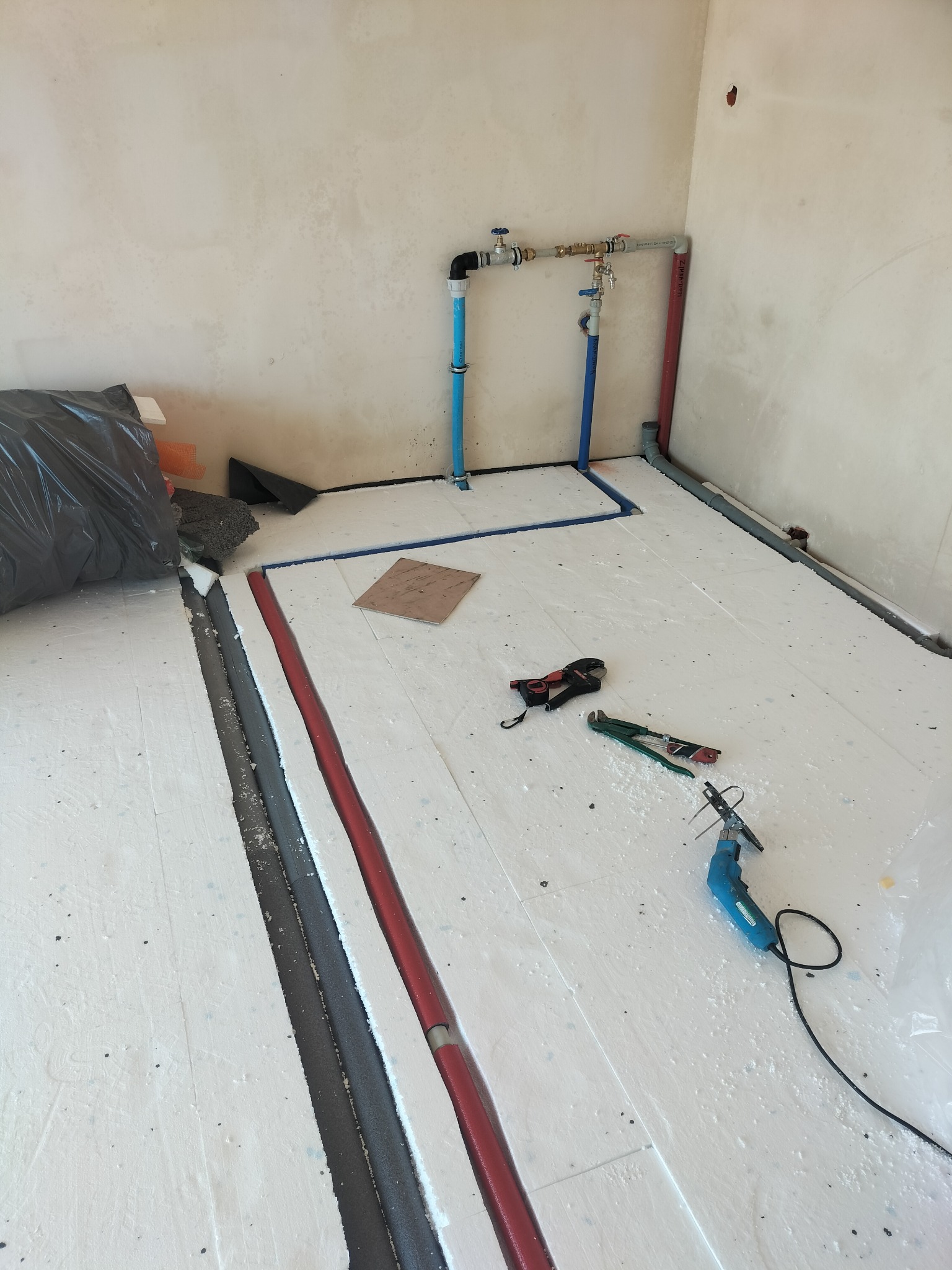 Rozłożona izolacja pod ogrzewanie podłogowe z widocznymi rurami, narzędziami i elementami instalacji. W tle surowe ściany w trakcie prac wykończeniowych.