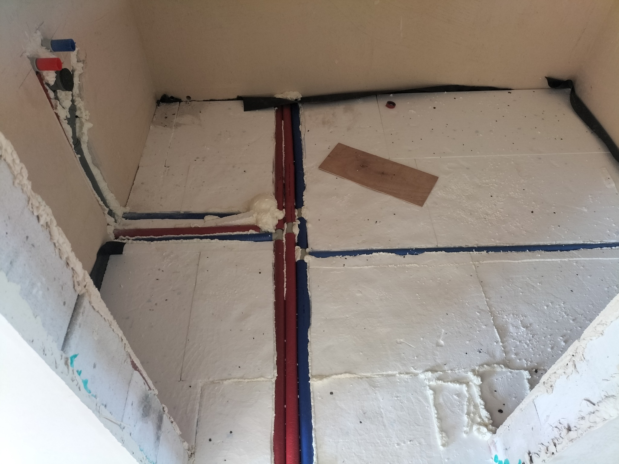 Izolacja podłogowa z rurami grzewczymi w kolorze czerwonym i niebieskim, zabezpieczonymi pianką montażową. Widoczna płyta drewniana na izolacji.